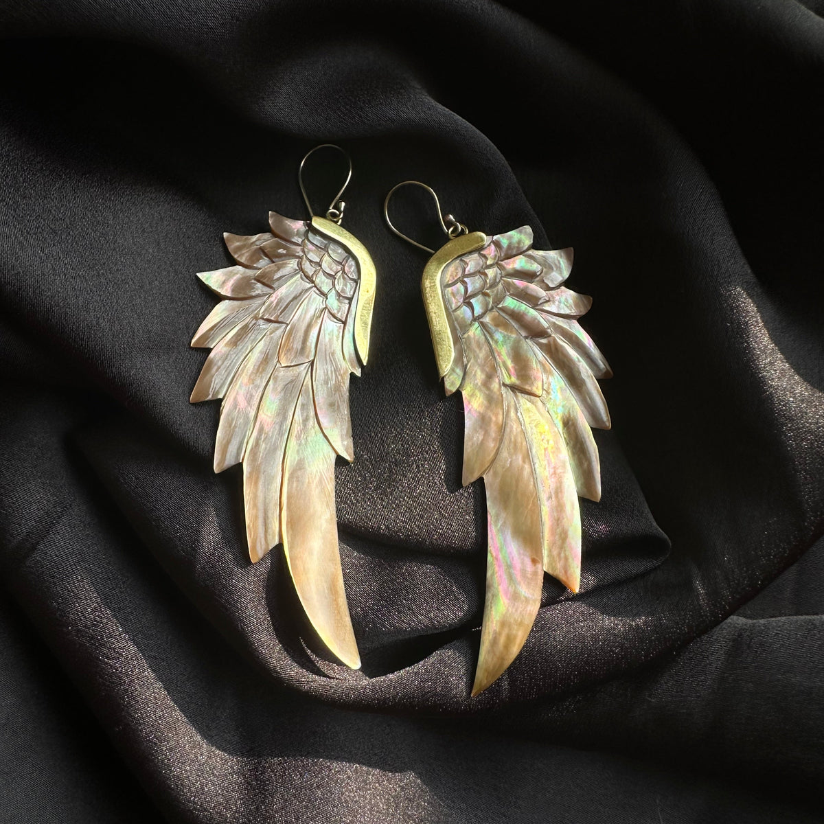 Pair of large statement drop earrings shaped like feathered angel wings, handcrafted from brass with iridescent mother-of-pearl inlay, displayed on dark silky fabric - Let's Jam jewellery collection