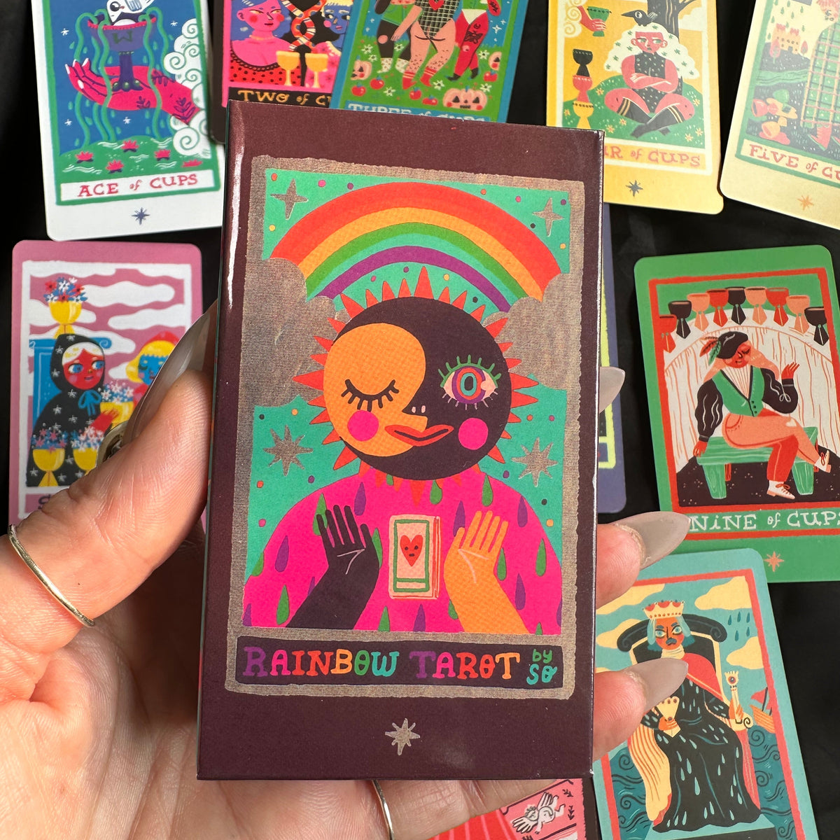 Hand holding "The Rainbow Tarot" deck box, featuring a colourful sun-and-rainbow illustration, displayed in front of a fanned spread of vividly illustrated tarot cards - Let's Jam collection