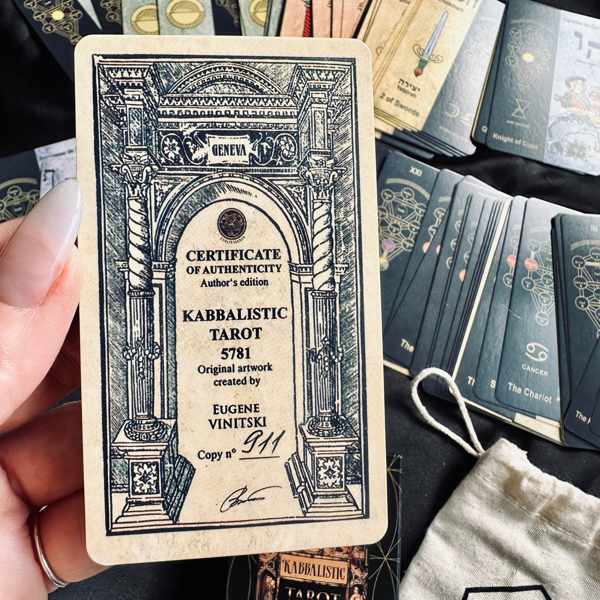 Hand holding the "Kabbalistic Tarot 5781" certificate of authenticity card in front of a fanned spread of Hebrew-lettered tarot cards, with the branded deck box and drawstring bag visible below - Let's Jam collection