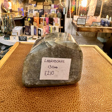 Load image into Gallery viewer, A large raw labradorite crystal stone with a handwritten price tag reading 'LABRADORITE 1311kg £210' sits on a textured brown leather surface inside a shop.