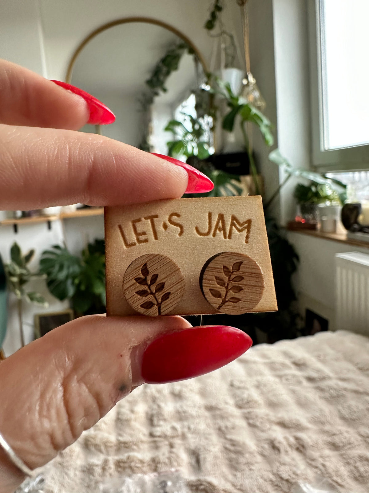 Hand holding a Let's Jam branded card with a pair of round wooden stud earrings engraved with a single leaf design, plants in the background - Let's Jam jewellery collection