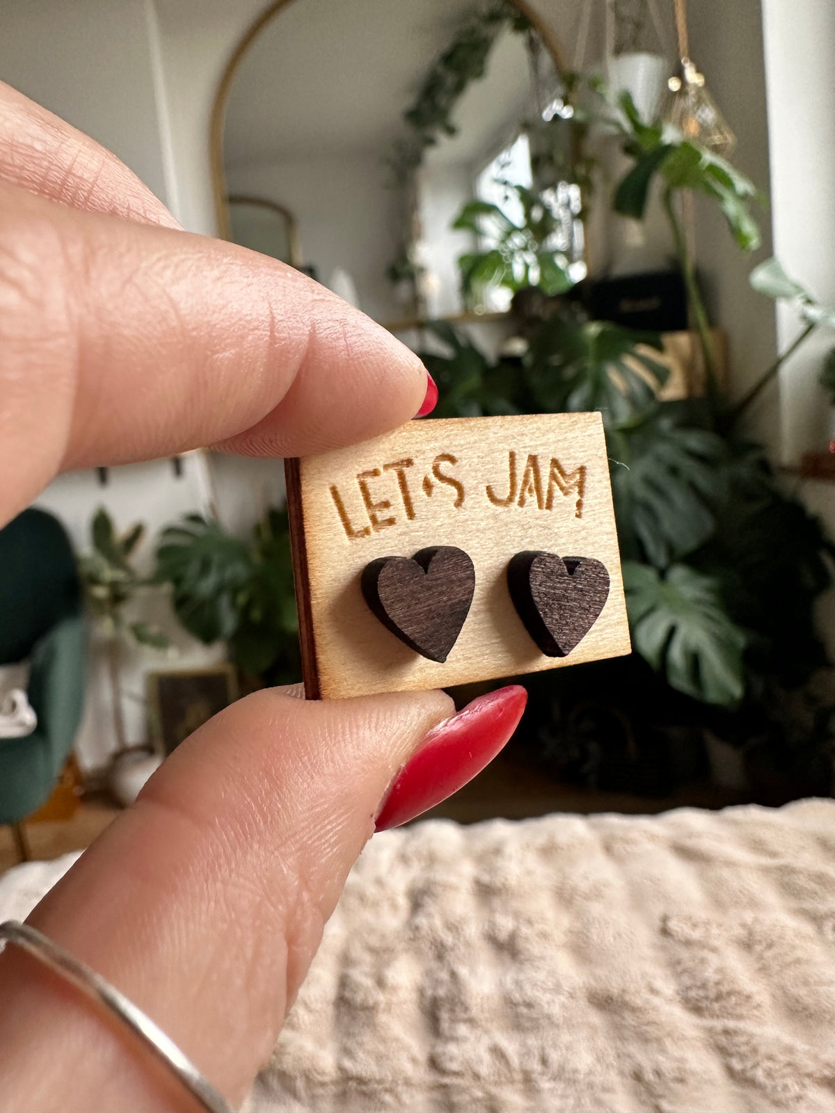 Hand holding a Let's Jam branded card with a pair of wooden stud earrings shaped like hearts, plants and a mirror in the background - Let's Jam jewellery collection