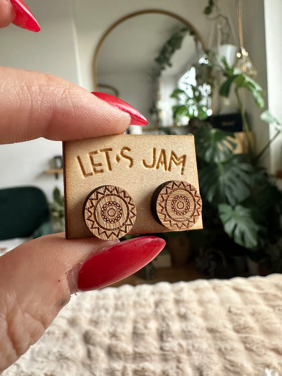 Hand holding a Let's Jam branded card with a pair of round wooden stud earrings engraved with an intricate mandala design, plants in the background - Let's Jam jewellery collection