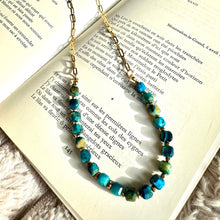 Load image into Gallery viewer, Blue tiger's eye beaded necklace with 18k gold-plated stainless steel chain, draped over an open book on a soft fabric background - Let's Jam jewellery collection