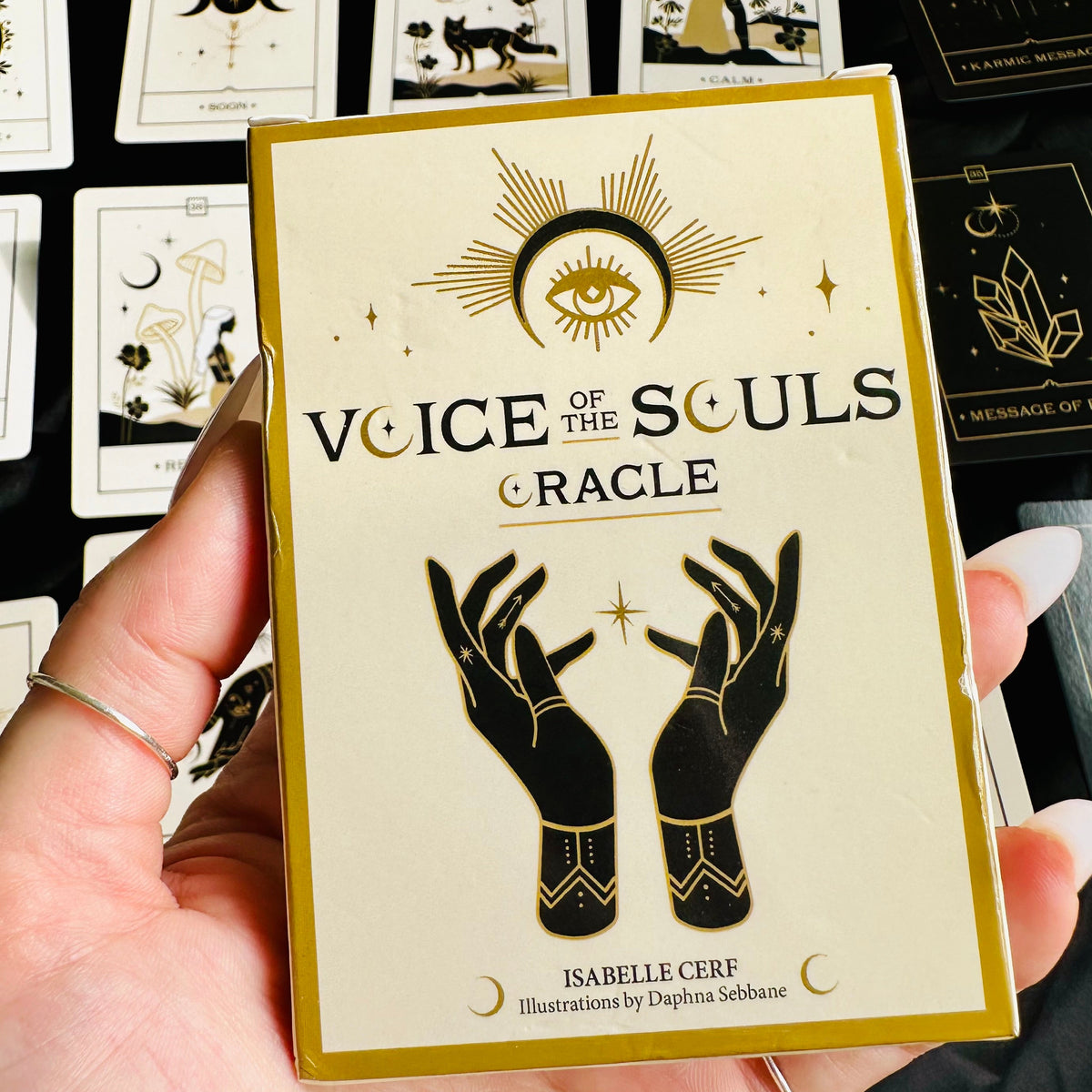 Hand holding the "Voice of the Souls Oracle" deck box, cream and gold with an eye and clasped-hands illustration, in front of a spread of matching black-and-gold oracle cards - Let's Jam collection