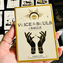 Load image into Gallery viewer, Hand holding the "Voice of the Souls Oracle" deck box, cream and gold with an eye and clasped-hands illustration, in front of a spread of matching black-and-gold oracle cards - Let's Jam collection