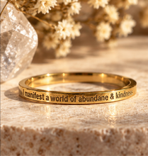 Load image into Gallery viewer, A polished gold ring engraved with the quote 'I manifest a world of abundance & kindness', in a warm, golden-lit setting on a textured stone surface with dried flowers and a crystal in the background.