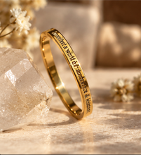 Load image into Gallery viewer, A gold band ring engraved with the phrase 'I manifest a world of abundance & kindness', leaning against a clear crystal on a warm beige surface with soft dried flowers in the background.