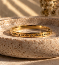 Load image into Gallery viewer, A polished gold ring engraved with the phrase 'I manifest a world of abundance & kindness' in black lettering, resting on a textured beige stone dish.