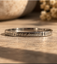 Load image into Gallery viewer, A polished silver ring engraved with the phrase 'I manifest a world of abundance & kindness' in a warm, rustic setting on a wooden surface.