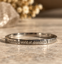 Load image into Gallery viewer, A polished silver ring engraved with the phrase 'I manifest a world of abundance & kindness' on a textured stone surface with a soft-focus background.