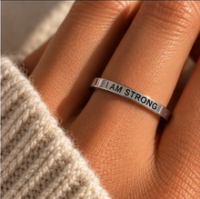 Load image into Gallery viewer, A close-up of a hand wearing a thin silver band ring engraved with the words 'I AM STRONG'. The ring is shown on a finger, with a beige knit sweater visible in the foreground and warm sunlight casting soft shadows.