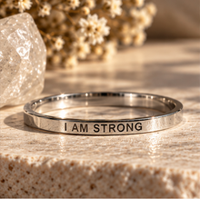Load image into Gallery viewer, A polished silver metal ring engraved with the words 'I AM STRONG' in black lettering, resting on a textured beige stone surface with dried flowers and a crystal in the background.