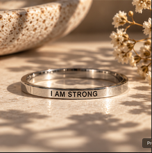 Load image into Gallery viewer, A polished silver-toned metal ring engraved with the words 'I AM STRONG' in black lettering, resting on a textured beige surface with soft shadows and dried flowers in the background.
