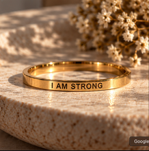 Load image into Gallery viewer, A gold-toned ring engraved with the words 'I AM STRONG' in black capital letters, resting on a textured beige stone surface with dried flowers in the background.