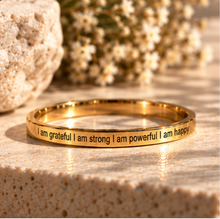 Load image into Gallery viewer, A gold-plated ring engraved with the affirmation 'I am grateful I am strong I am powerful I am happy' in black lettering. The ring is displayed on a textured beige stone surface with a blurred background of a rock and dried flowers.