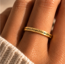 Load image into Gallery viewer, A close-up of a gold band ring worn on a finger, featuring the engraved text 'Everything changes & this too shall pass'. The ring is shown on a person's finger with a cream-colored knit sweater visible at the wrist, set against a warm, softly blurred background.