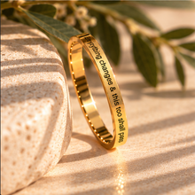 Load image into Gallery viewer, A polished gold ring engraved with the quote 'Everything changes & this too shall pass' in black lettering, displayed on a textured beige stone with green leaves in warm sunlight.
