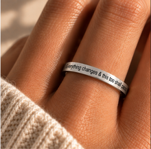 Load image into Gallery viewer, A close-up of a hand wearing a slim silver band ring with engraved black text that reads, 'Everything changes & this too shall pass.' The photo is shot in warm natural light, with a cream-colored knit sleeve visible at the bottom left.