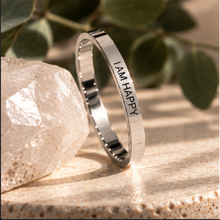 Load image into Gallery viewer, A polished silver band ring engraved with the words 'I AM HAPPY' in black lettering, standing upright on a textured beige stone surface next to a faceted crystal, with soft-focus green leaves in the background.