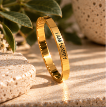Load image into Gallery viewer, A polished gold ring with the words 'I AM HAPPY' engraved vertically, standing upright on a textured beige stone surface with soft-focus green leaves and a ceramic pot in the background.