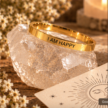 Load image into Gallery viewer, A polished gold ring engraved with 'I AM HAPPY' in black lettering, displayed on a faceted clear crystal cluster. The setting includes warm bokeh background elements, dried white flowers, and a sun-face card.