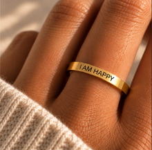 Load image into Gallery viewer, A close-up of a hand wearing a slim gold band ring engraved with the words 'I AM HAPPY' in black lettering, styled with a cream knit sweater cuff.