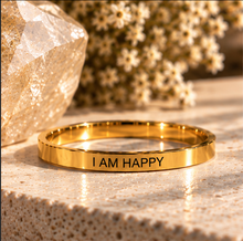 Load image into Gallery viewer, A polished gold ring engraved with the words 'I AM HAPPY' in black lettering, resting on a textured beige stone surface with warm, sunlit dried flowers and a faceted crystal in the background.