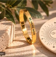 Load image into Gallery viewer, A polished gold ring with the words 'BE HERE NOW' engraved in black, displayed on a textured beige surface with decorative leaves and a stone block.