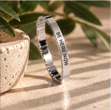 Load image into Gallery viewer, A polished silver-toned metal ring with the words 'BE HERE NOW' engraved vertically, displayed on a textured beige surface with soft shadows and green leaves in the background.