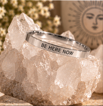 Load image into Gallery viewer, A polished silver ring engraved with the words 'BE HERE NOW' in black capital letters, displayed on a clear quartz crystal cluster with a warm, bohemian background.