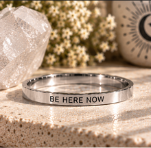 Load image into Gallery viewer, A polished silver-toned metal ring engraved with the words 'BE HERE NOW' in black capital letters, resting on a textured beige stone surface. The background is softly blurred with a crystal, small white flowers, and a decorative ceramic vase.