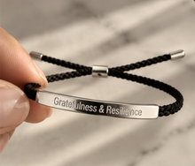 Load image into Gallery viewer, Hand holding a black cord bracelet with a silver Gratefulness and Resilience bar - Let's Jam jewellery, Camden Town