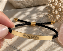 Load image into Gallery viewer, A close-up of a hand holding a black braided cord bracelet with a polished gold-tone metal bar engraved with the phrase 'You've got this'. The bracelet features a gold sliding knot and a gold spacer bead, presented on a light marble surface with soft-focus dried flowers in the background.
