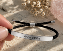 Load image into Gallery viewer, A close-up of a hand holding a black braided cord bracelet with a polished silver bar engraved with the words 'You've got this'. The background features a textured stone surface with dried flowers and a pink crystal.