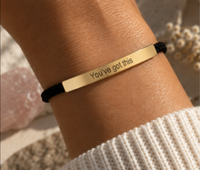 Load image into Gallery viewer, A close-up of a person's wrist wearing a gold bar bracelet engraved with the words 'You've got this' on a black cord, with a white knit sleeve visible at the bottom.