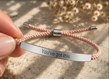 Load image into Gallery viewer, A close-up of a hand holding a pink braided cord bracelet with a polished silver metal bar engraved with the words 'You've got this'. The background is a textured beige stone surface with soft-focus dried flowers.