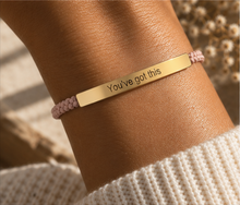 Load image into Gallery viewer, A close-up of a person's wrist wearing a gold bar bracelet engraved with the words 'You've got this' in black lettering, paired with a light pink braided cord bracelet, set against a warm, sunlit background.