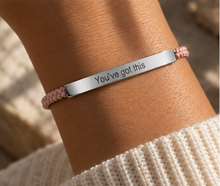 Load image into Gallery viewer, A close-up of a person's wrist wearing a silver bar bracelet engraved with the words 'You've got this', paired with a light pink braided cord bracelet, set against a soft beige knit sweater and blurred natural background.