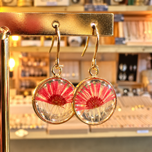Load image into Gallery viewer, Round resin earrings with a bright red pressed flower hanging on a gold stand in the shop - Let's Jam, Camden Market, London