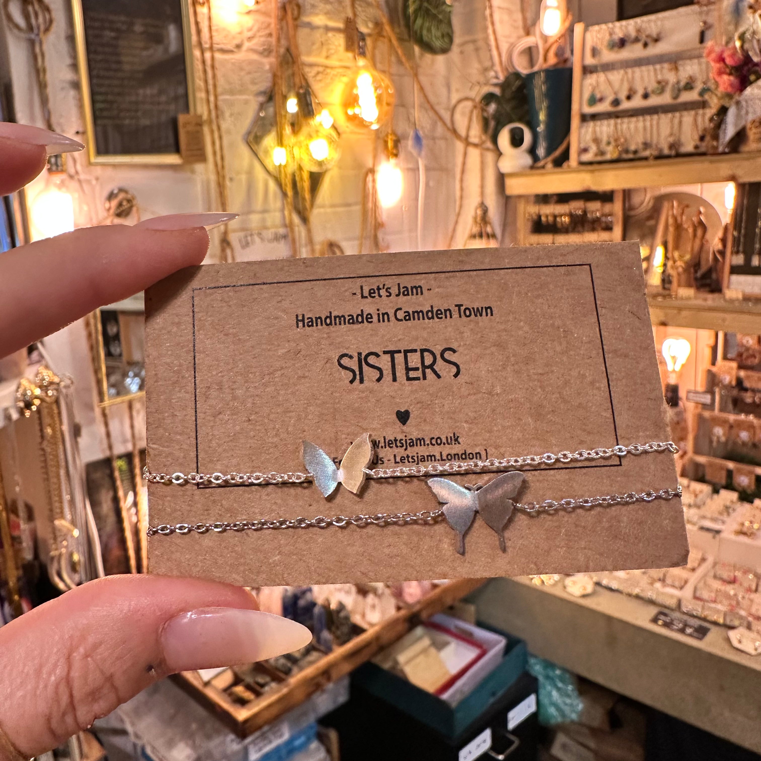 A hand holds a brown kraft display card featuring two delicate silver chain bracelets with butterfly charms. The card is printed with the brand name 'Let's Jam' and the style name 'SISTERS'.