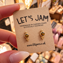 Load image into Gallery viewer, Gold stud earrings with small tiger's eye stones on a branded card held in the hand - Let's Jam jewellery, Camden Market