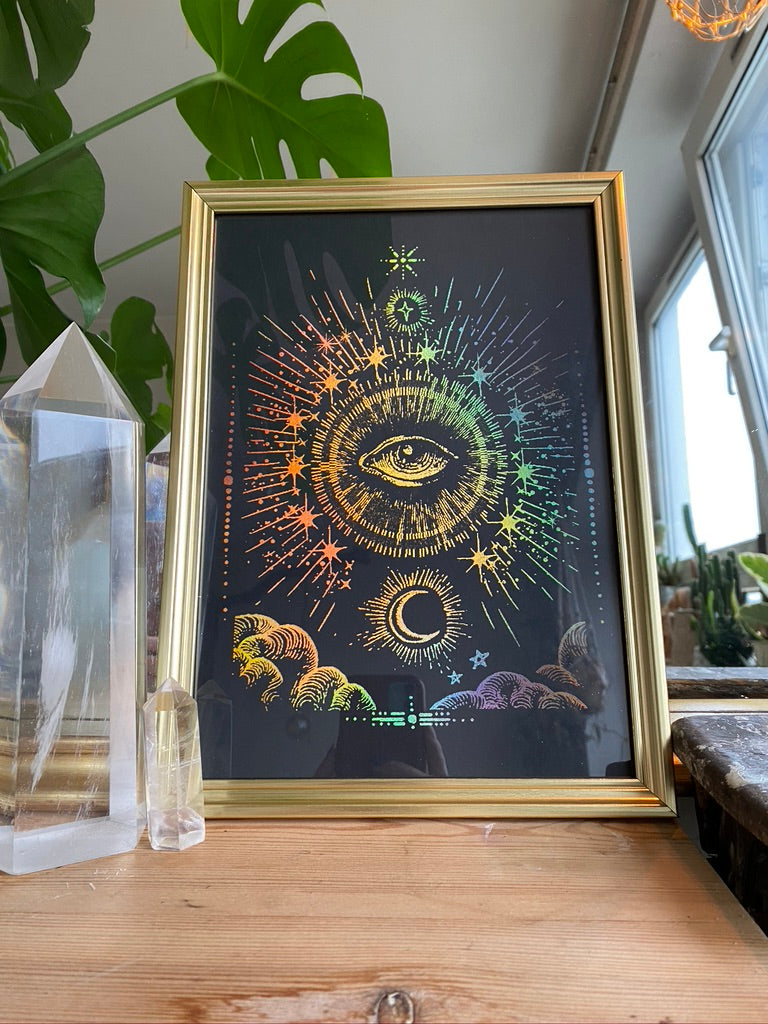 A gold-framed rainbow holographic celestial print of an all-seeing eye with sunburst rays, stars, and a crescent moon, displayed on a wooden table beside crystal points - Let's Jam print collection
