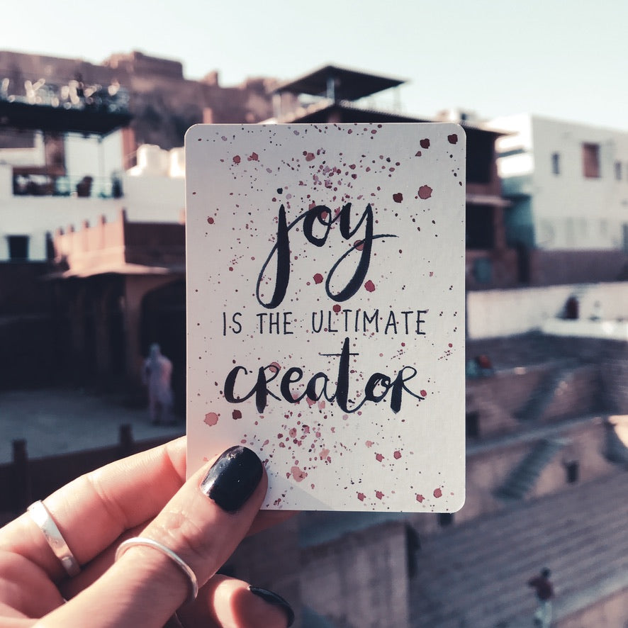 Hand holding a tarot card reading "Joy is the ultimate creator" against a rooftop cityscape - Let's Jam collection