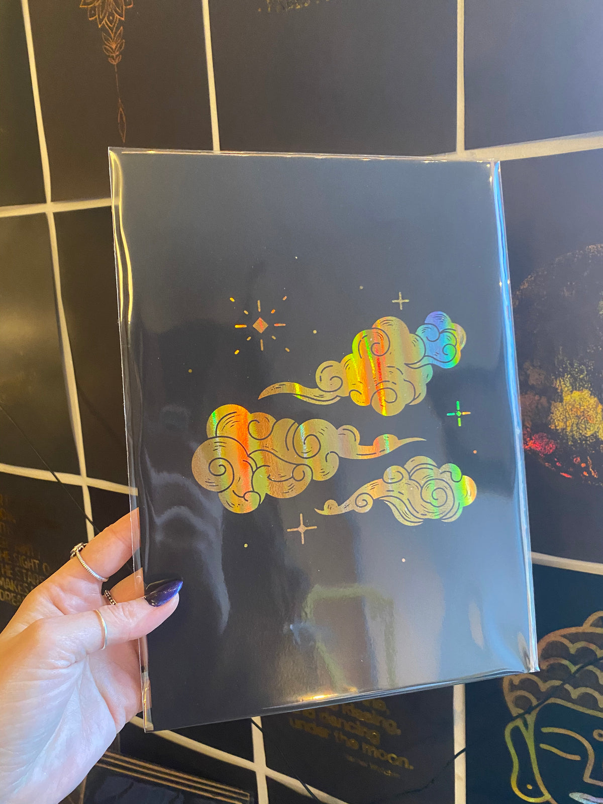 A hand holding a rainbow holographic A4 print of decorative swirling clouds and stars, against a gold-tiled wall - Let's Jam print collection