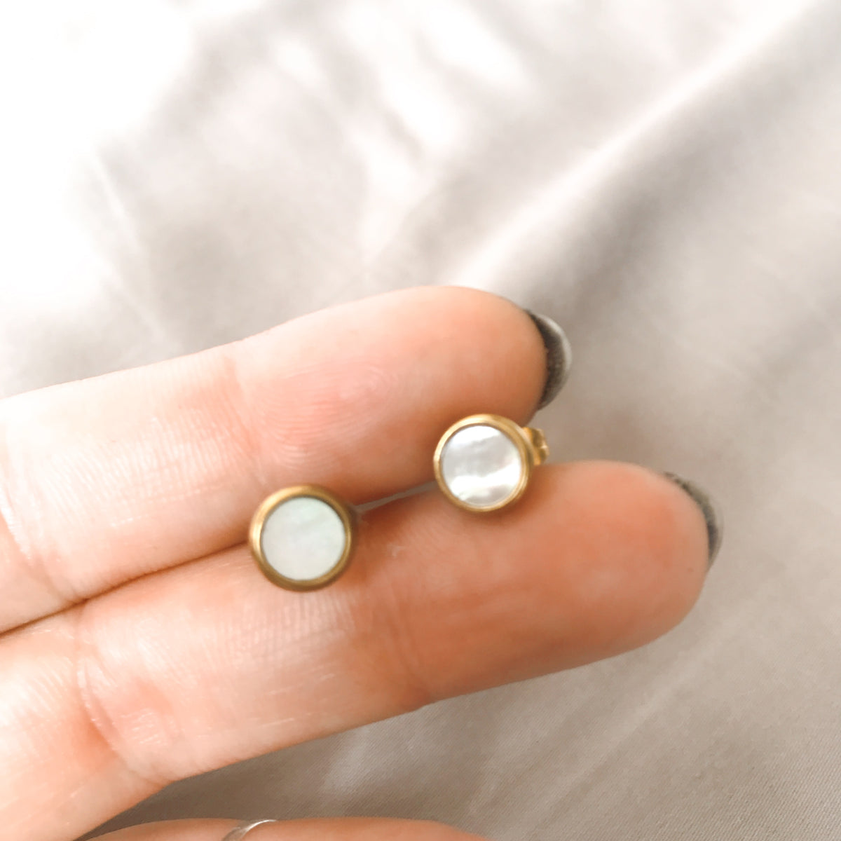 Two round mother-of-pearl stud earrings in a gold-tone stainless steel setting, shown held between fingers to show their soft iridescent shimmer - Let's Jam jewellery collection