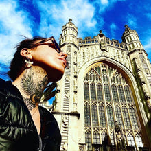 Load image into Gallery viewer, Model wearing gold-plated Isis wing drop earrings, photographed outdoors against a Gothic cathedral facade - Let's Jam jewellery collection
