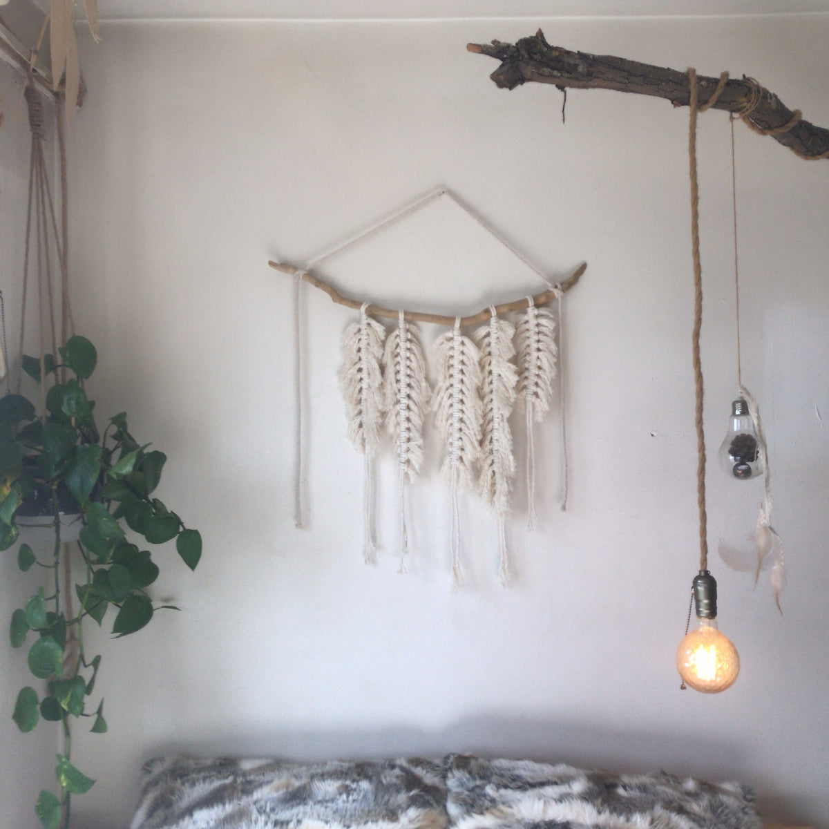 A handmade macrame wall hanging with feather-like fringed strands on a driftwood branch, styled on a bedroom wall beside a hanging plant and a glowing pendant bulb - Let's Jam collection
