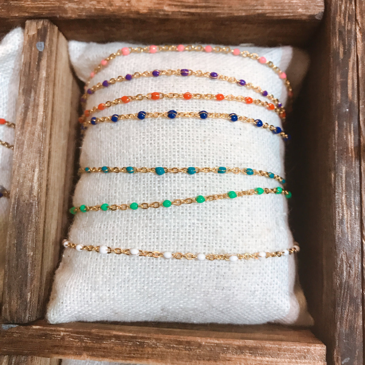 A row of dainty gold-tone chain bracelets with small beaded accents in a range of colours, arranged on a textured cushion inside a wooden box - Let's Jam jewellery collection