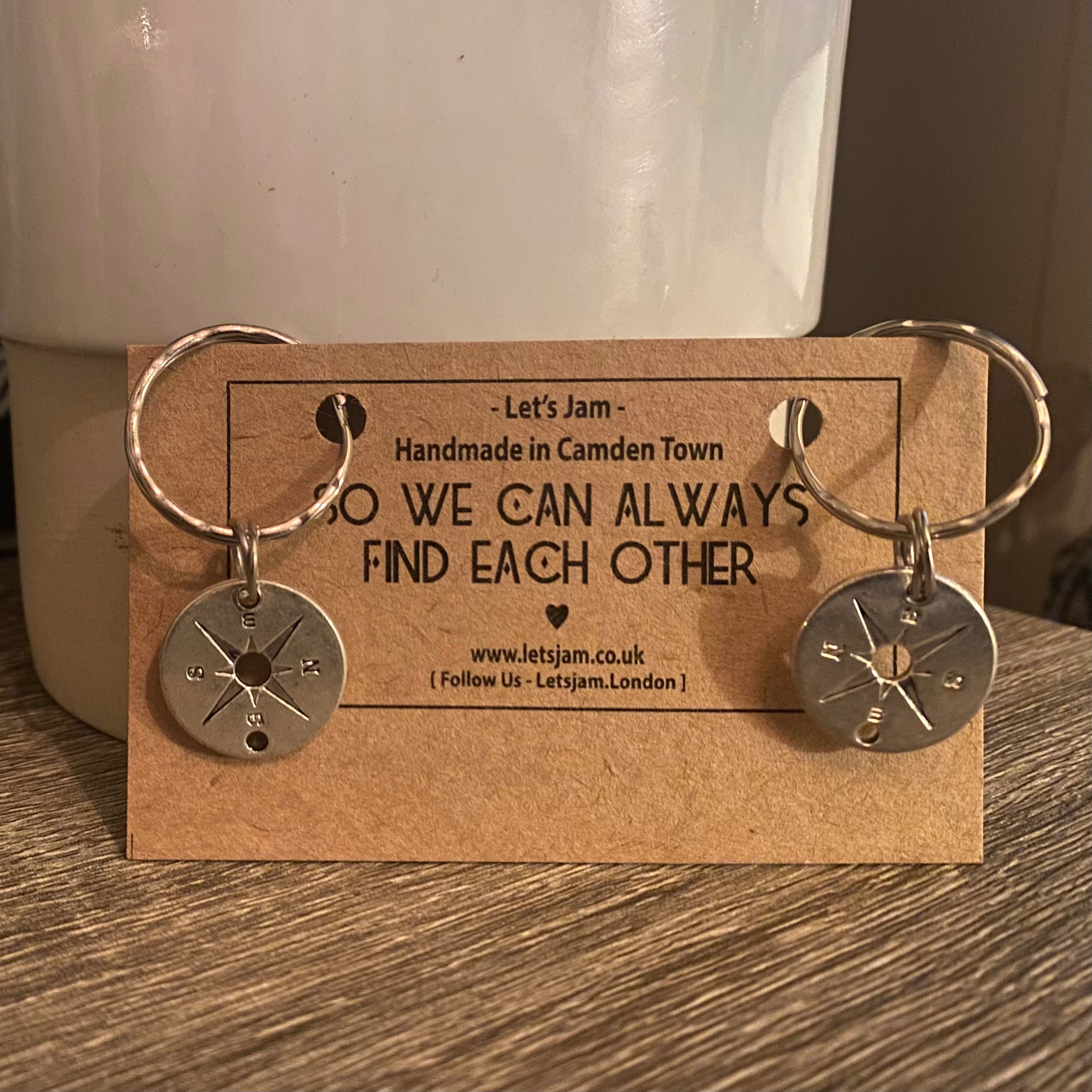 Two matching silver compass keyrings on a Let's Jam "So We Can Always Find Each Other" branded card, propped on a wooden table - Let's Jam collection